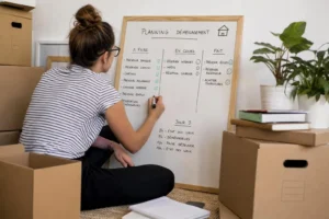 Une personne écrivant sur un tableau blanc intitulé “Planning déménagement” entourée de cartons et de plantes, planifiant les tâches. intégration: "comment organiser son déménagement" naturellement.
